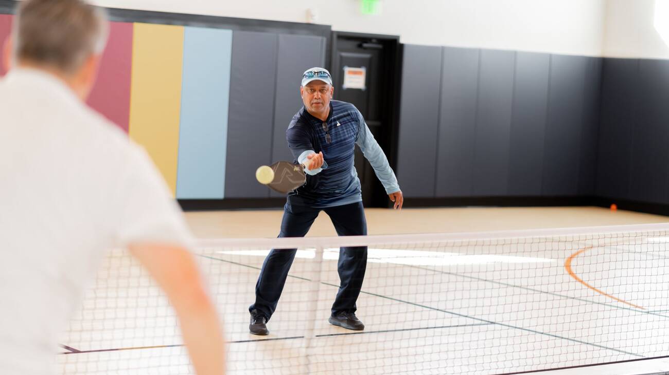 An older man returning a serve on an indoor pickleball court.
