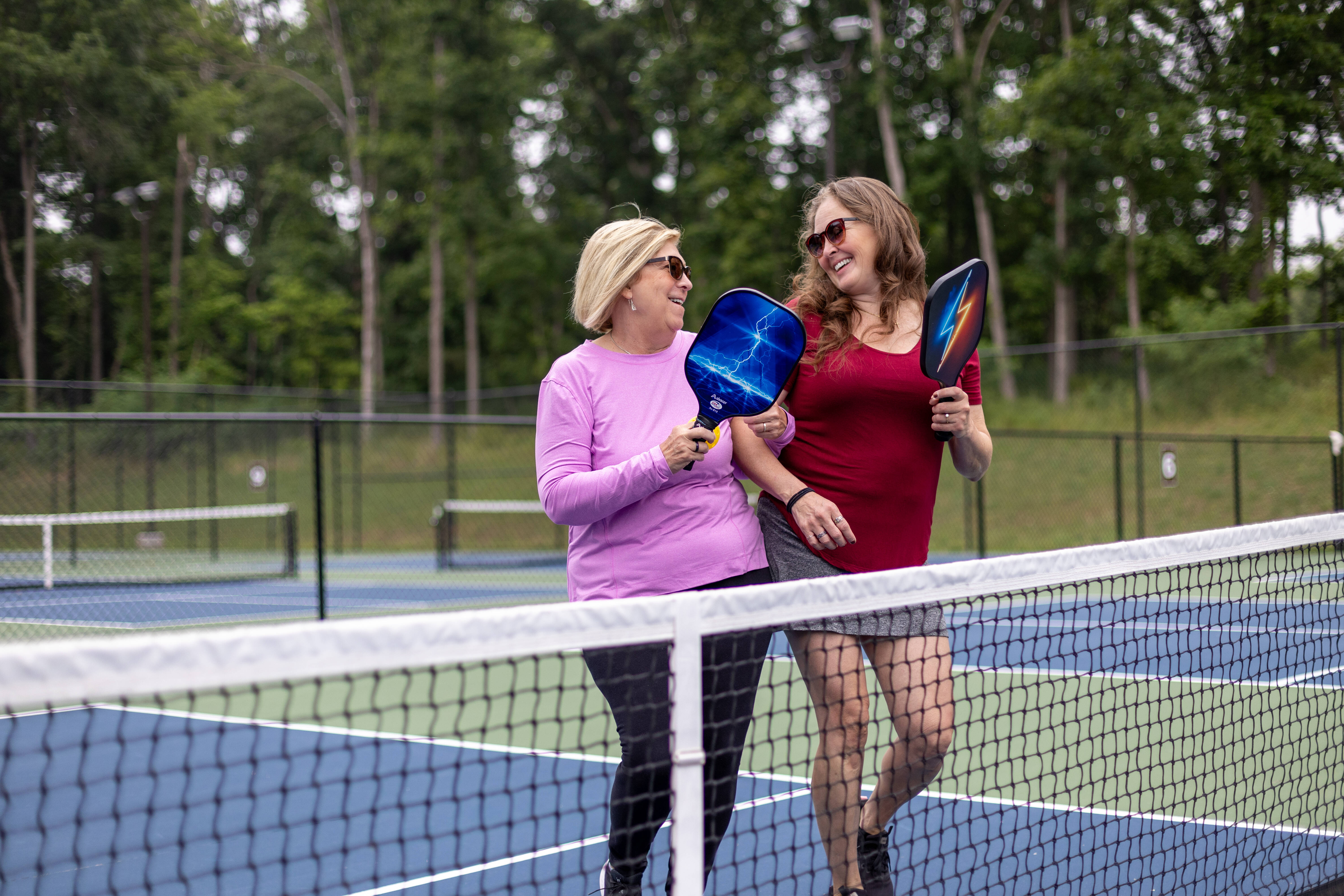 Two friends wearing sunglasses chatting as they play pickleball on an outdoor pickleball court.